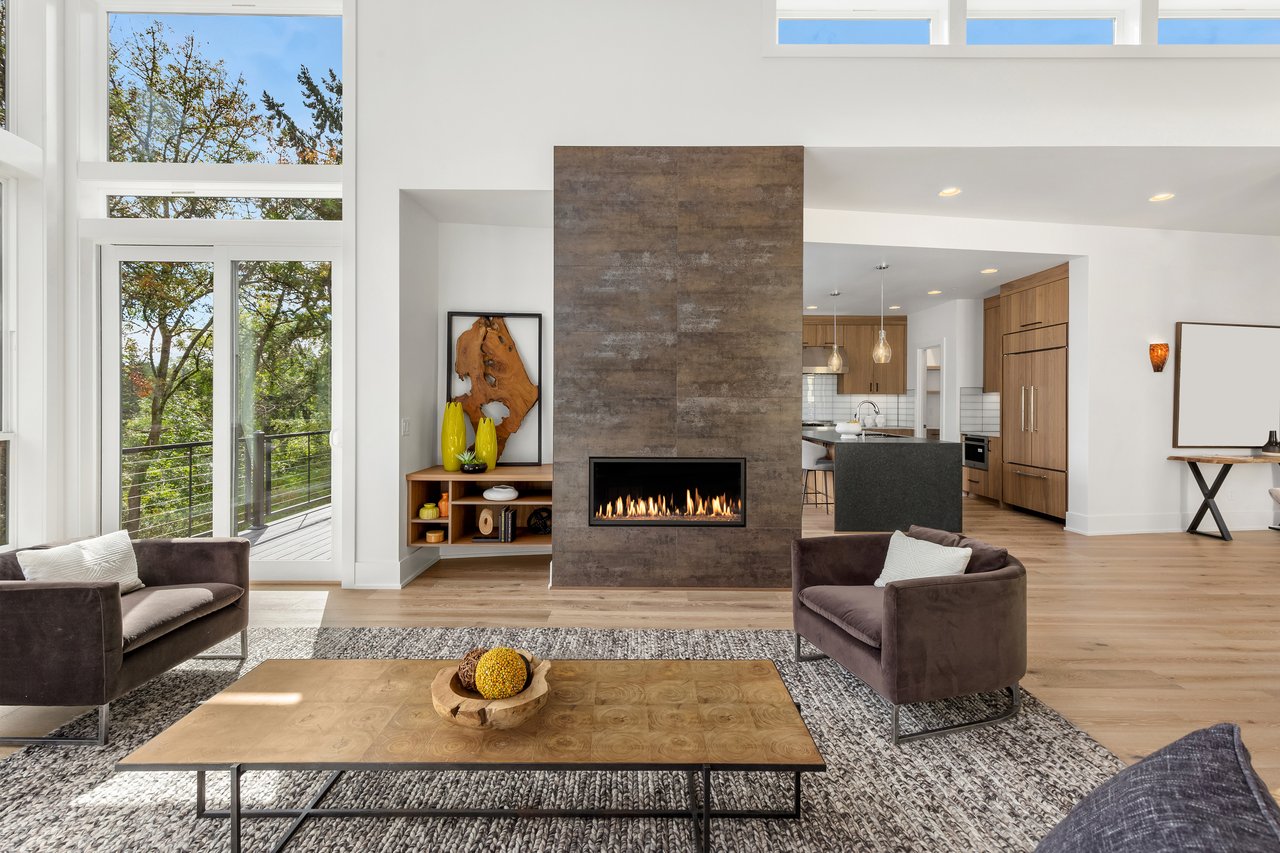 Open-plan living room with a tall stone fireplace, large windows, and a kitchen with wood cabinets.