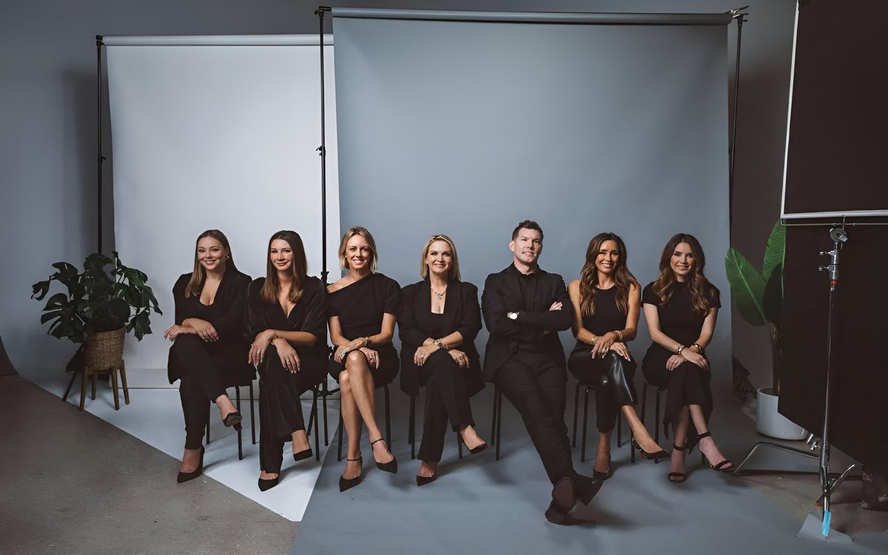  A team of six real estate professionals, three men and three women, sit on chairs in a photo studio against a grey backdrop.