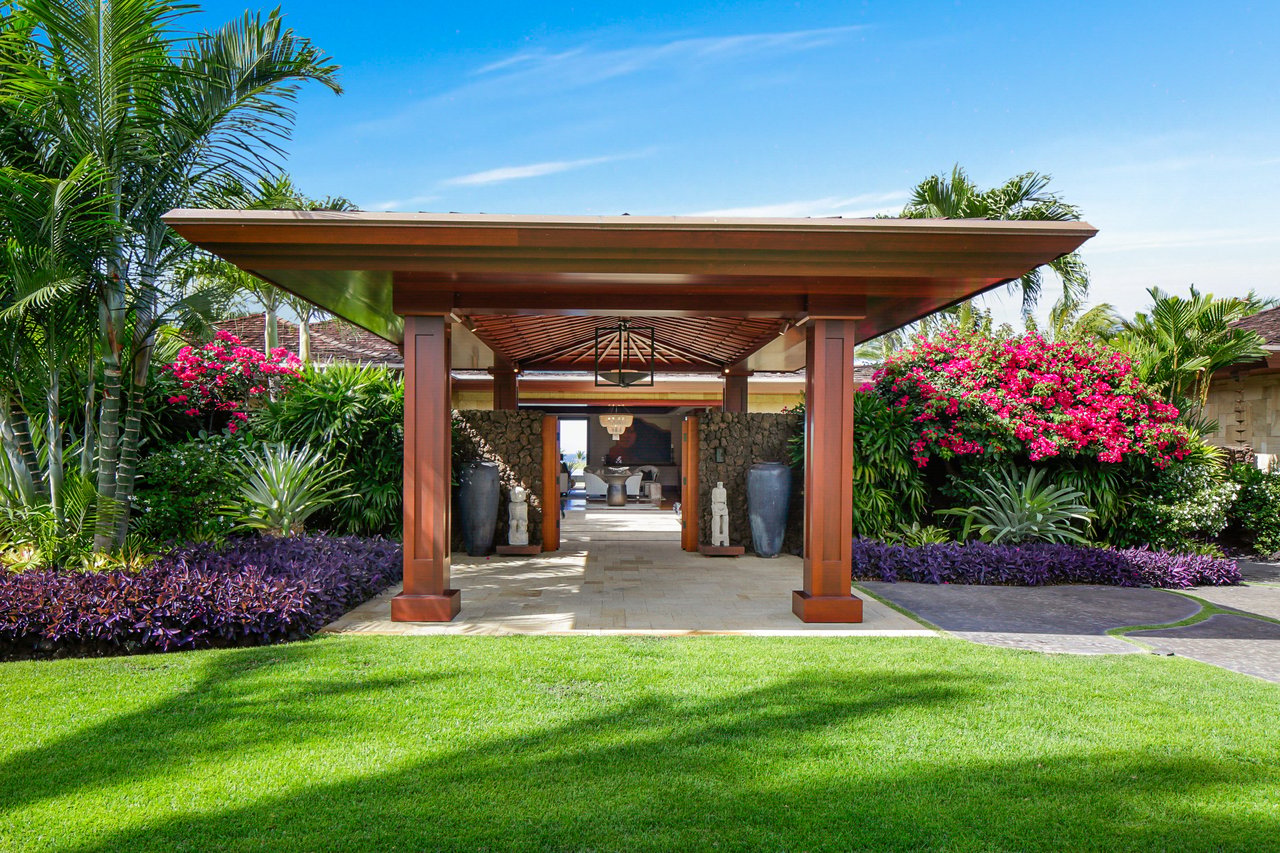Tropical property entrance framed by a broad wooden pavilion, palm trees, and pink flowering shrubs.