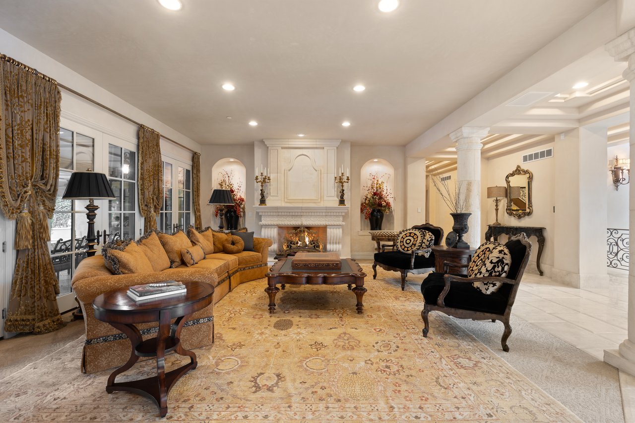Elegant living room with a carved fireplace, gold sofa, patterned armchairs, and large windows.