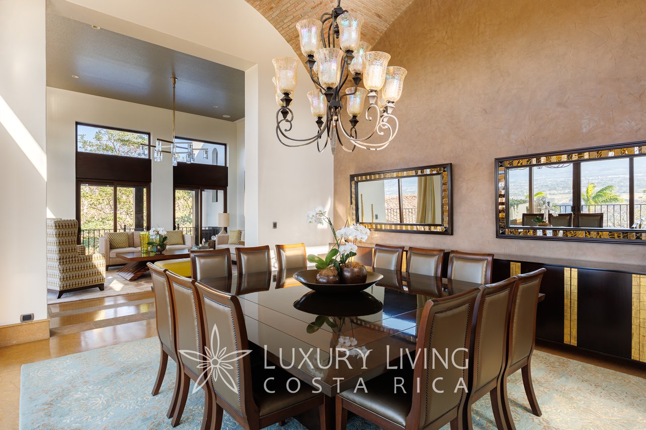 Dining room with a long dark table, upholstered chairs, chandelier, and tall windows overlooking a valley.