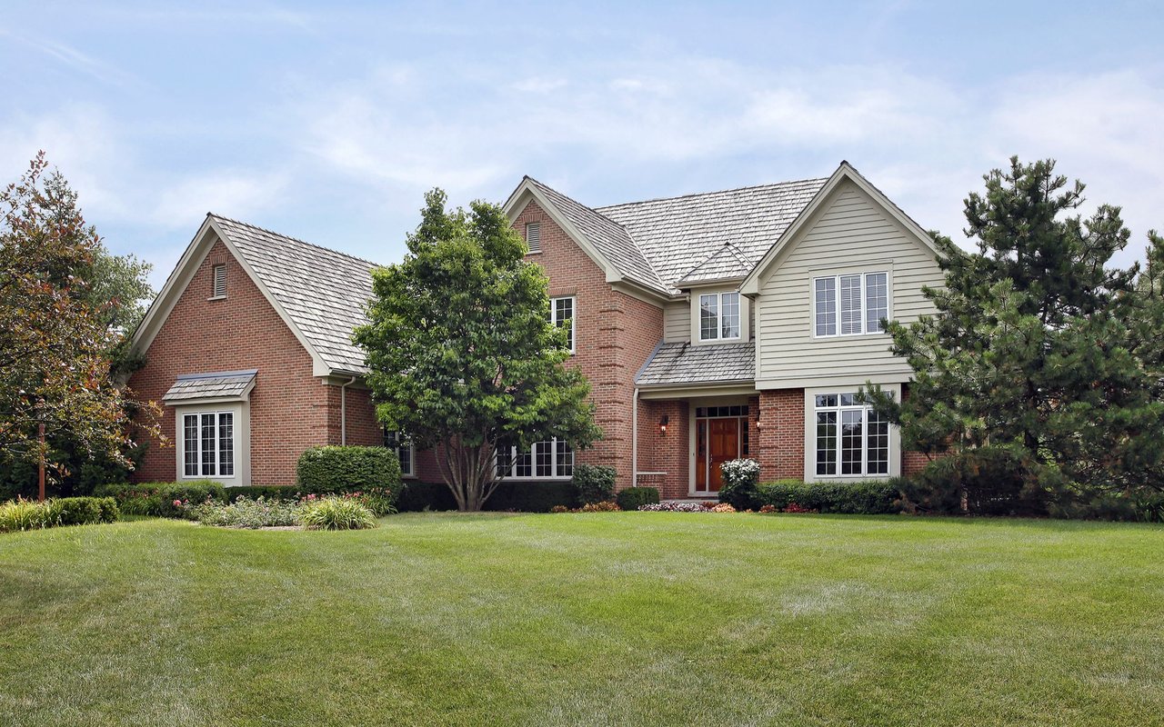 Two-story brick and light-sided home with gabled roofs, mature trees, and a broad lawn.