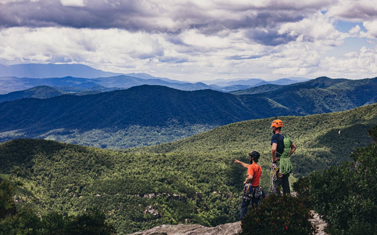 Two climbers survey forested mountain ridges beneath a cloudy sky.