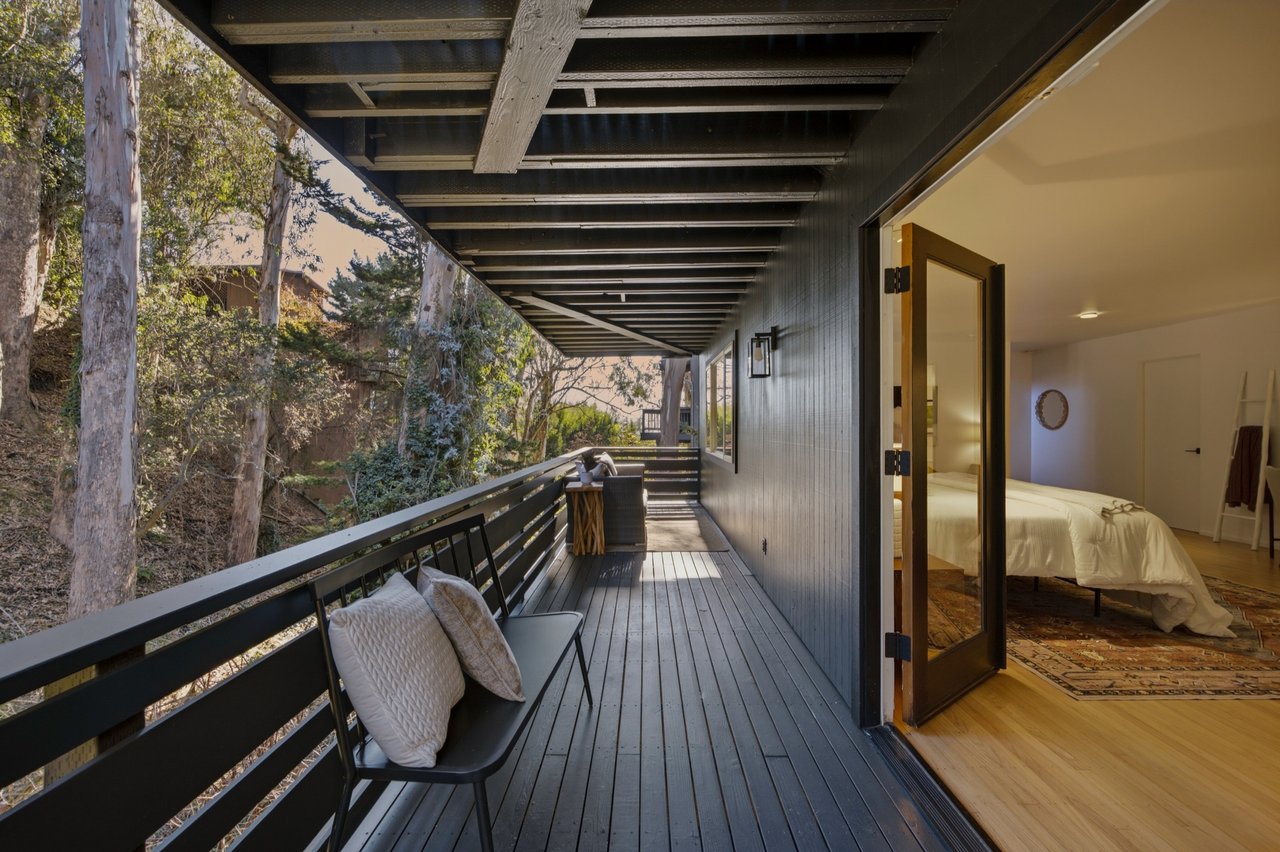 Covered deck with dark wood flooring and seating beside a bedroom opening onto a wooded hillside.