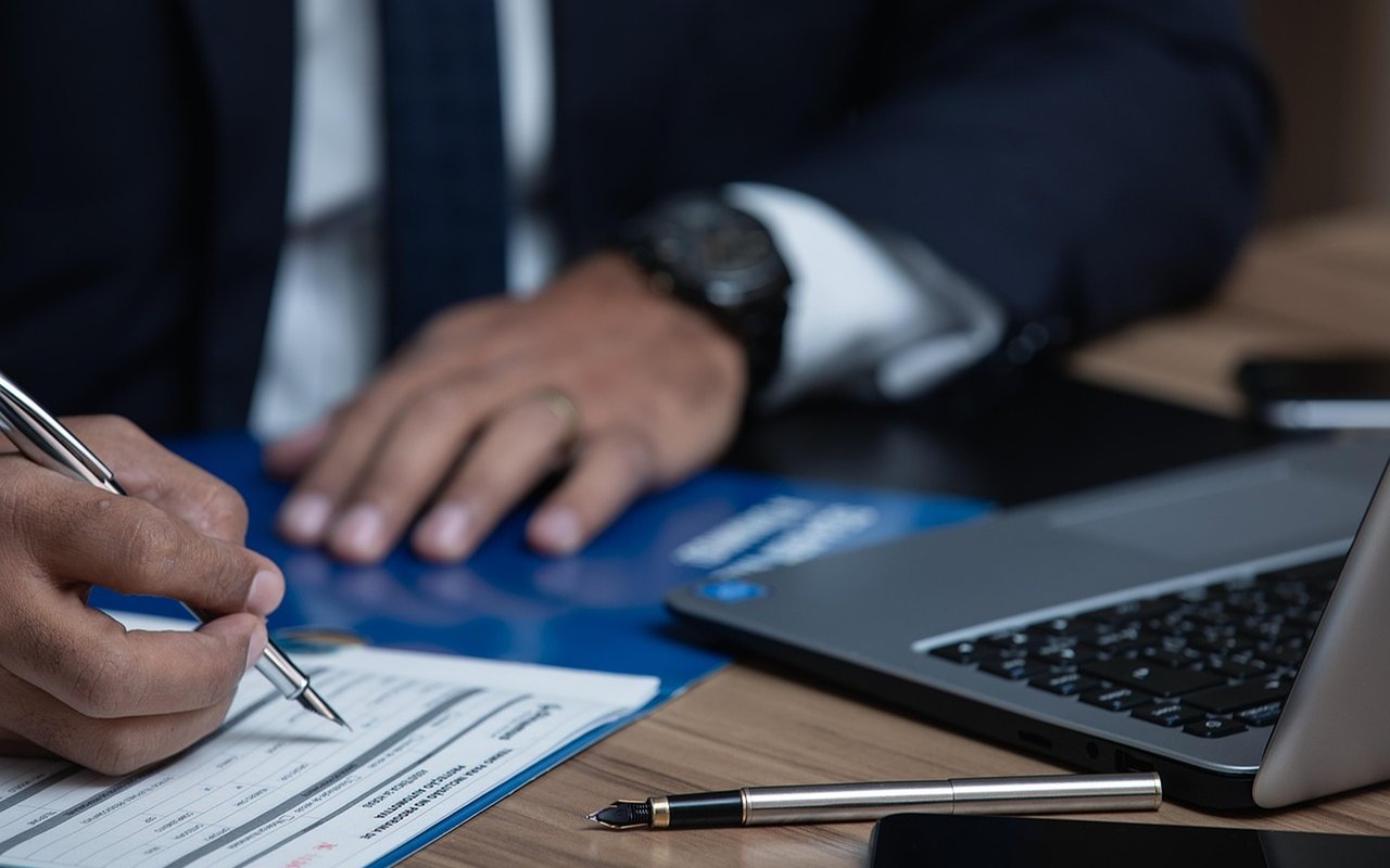 Hands fill out paperwork beside an open laptop.