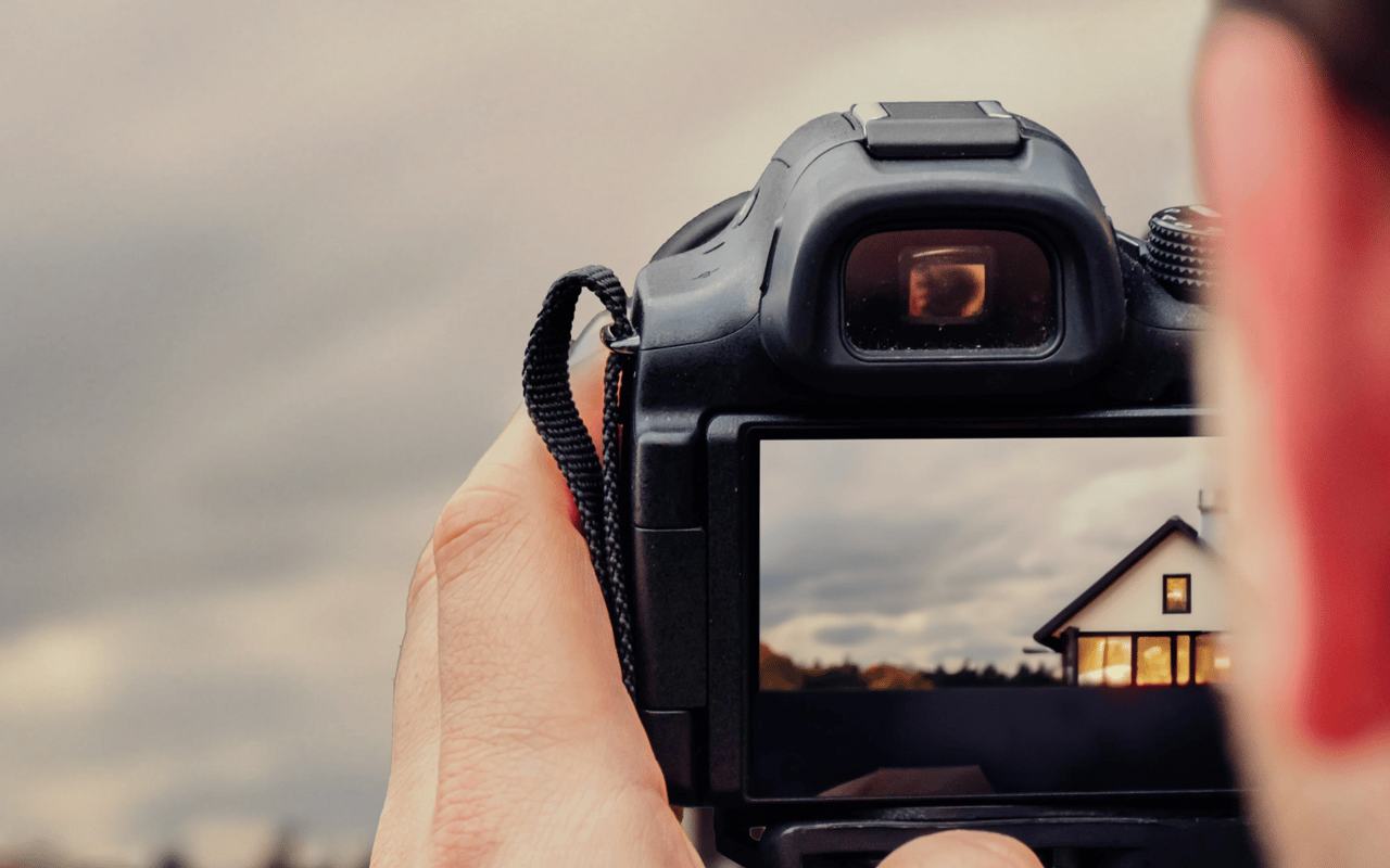 A person photographs a house with a digital camera, its windows glowing on the camera screen.