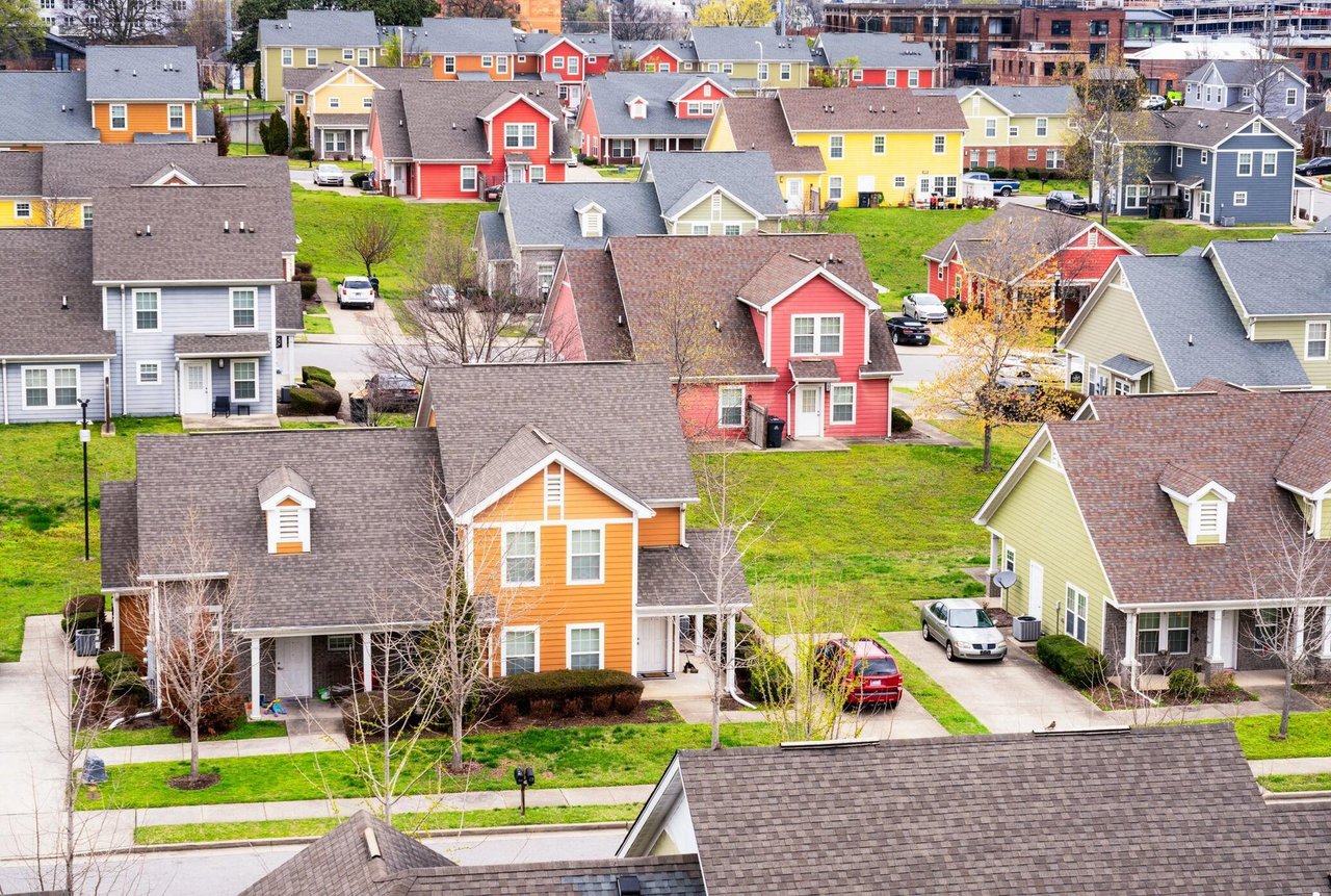 Colorful suburban neighborhood with vibrant houses in red, yellow, orange, and green, neatly arranged on a grassy landscape. The scene is peaceful and bright.