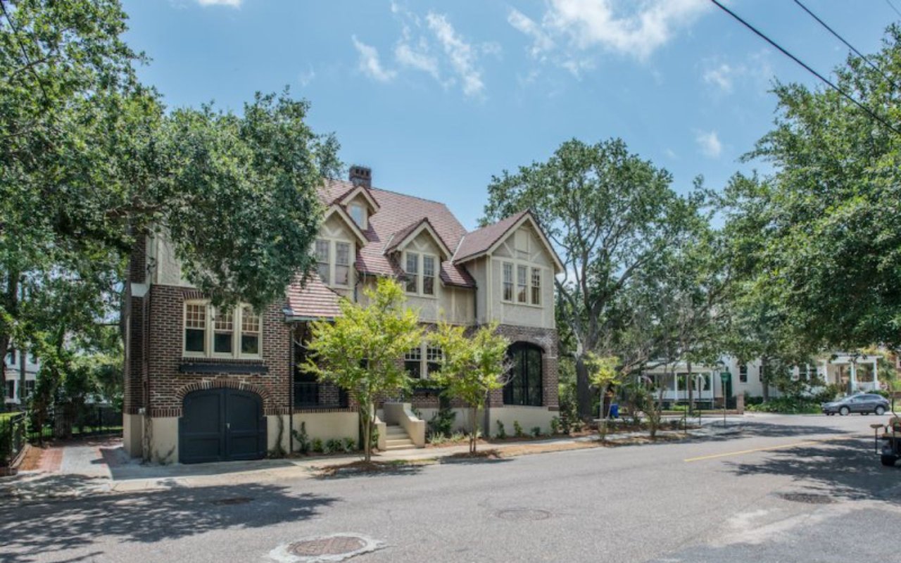 183 Tradd Street home with brick walls, a steep gabled roof, and dark garage doors beneath mature trees.