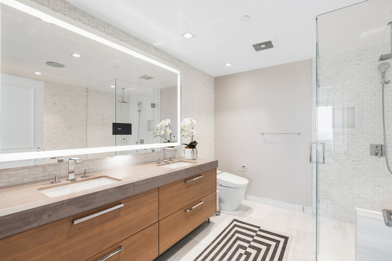 Modern bathroom with a double-sink wood vanity, illuminated mirror, glass shower, and geometric rug.