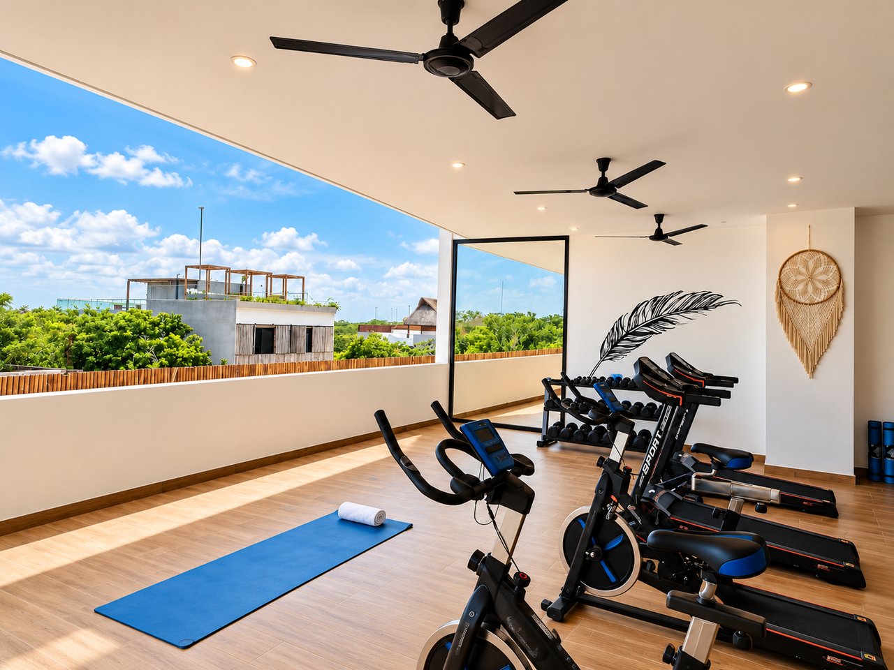 Fitness room with exercise bikes, treadmills, weights, and a yoga mat beside wide openings overlooking greenery.
