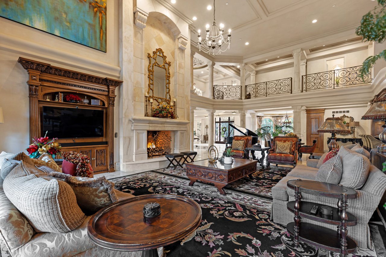 Grand living room with ornate fireplace, wrought-iron balcony, chandelier, and patterned rug.