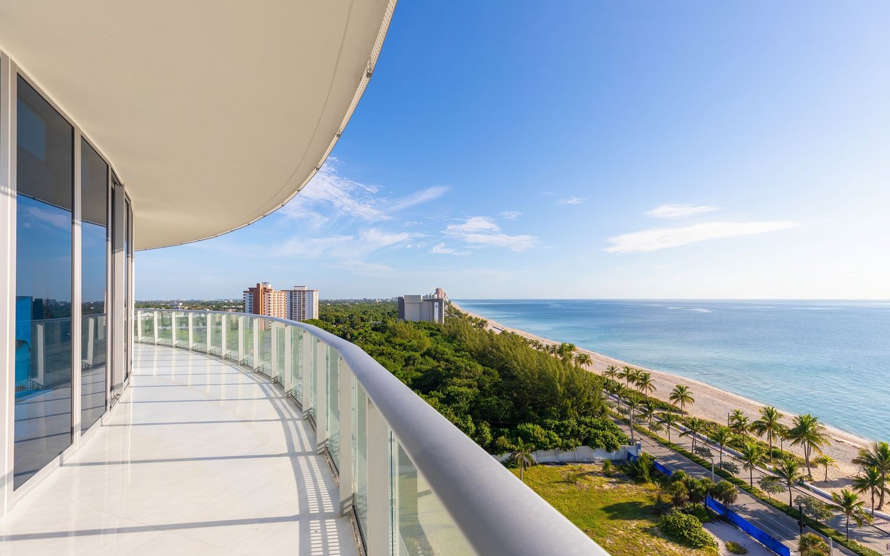 Curved glass-railed balcony overlooking a palm-lined beach and ocean.
