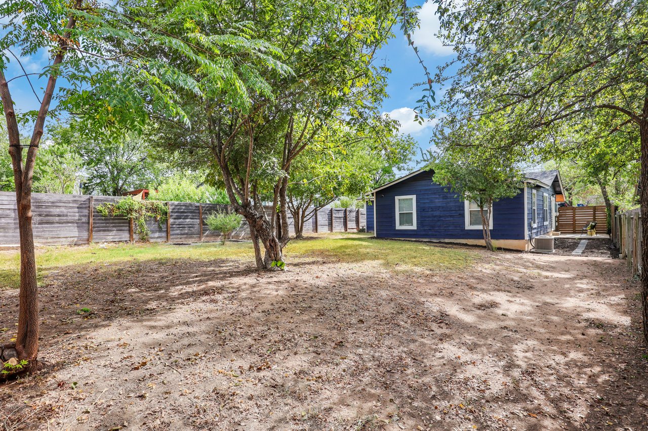 Tree-shaded backyard with a blue house, open yard, and wooden fencing.