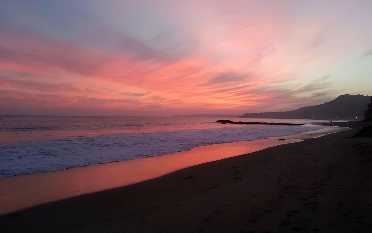 Beach at dusk with waves, a rocky outcrop, and a pink sunset over the distant coastline.