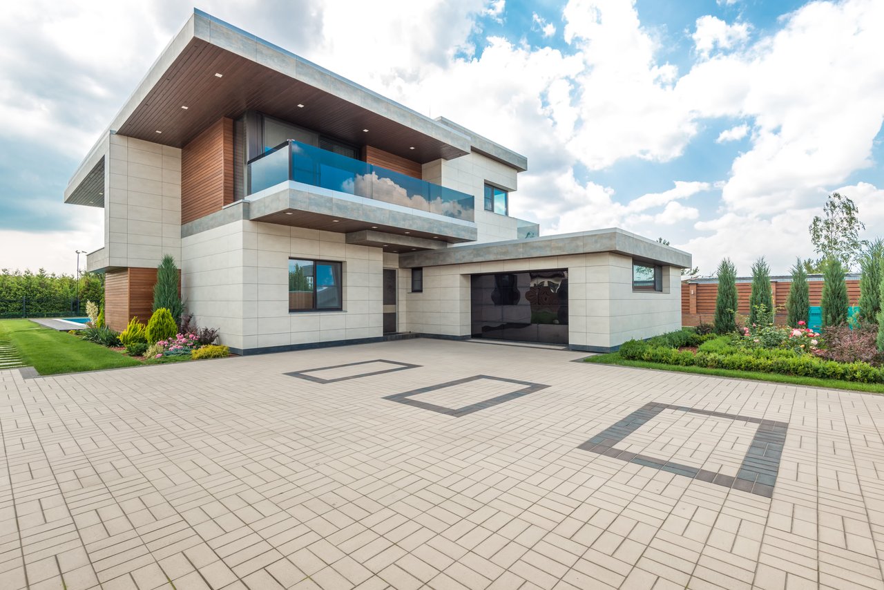 Modern home with a glass balcony, geometric-paved driveway, landscaped garden, and pool glimpse.