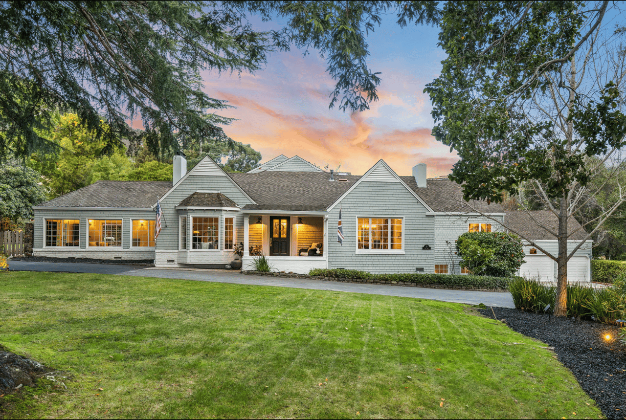 Gray-shingled home with multiple gables, warmly lit windows, and a broad lawn at dusk.