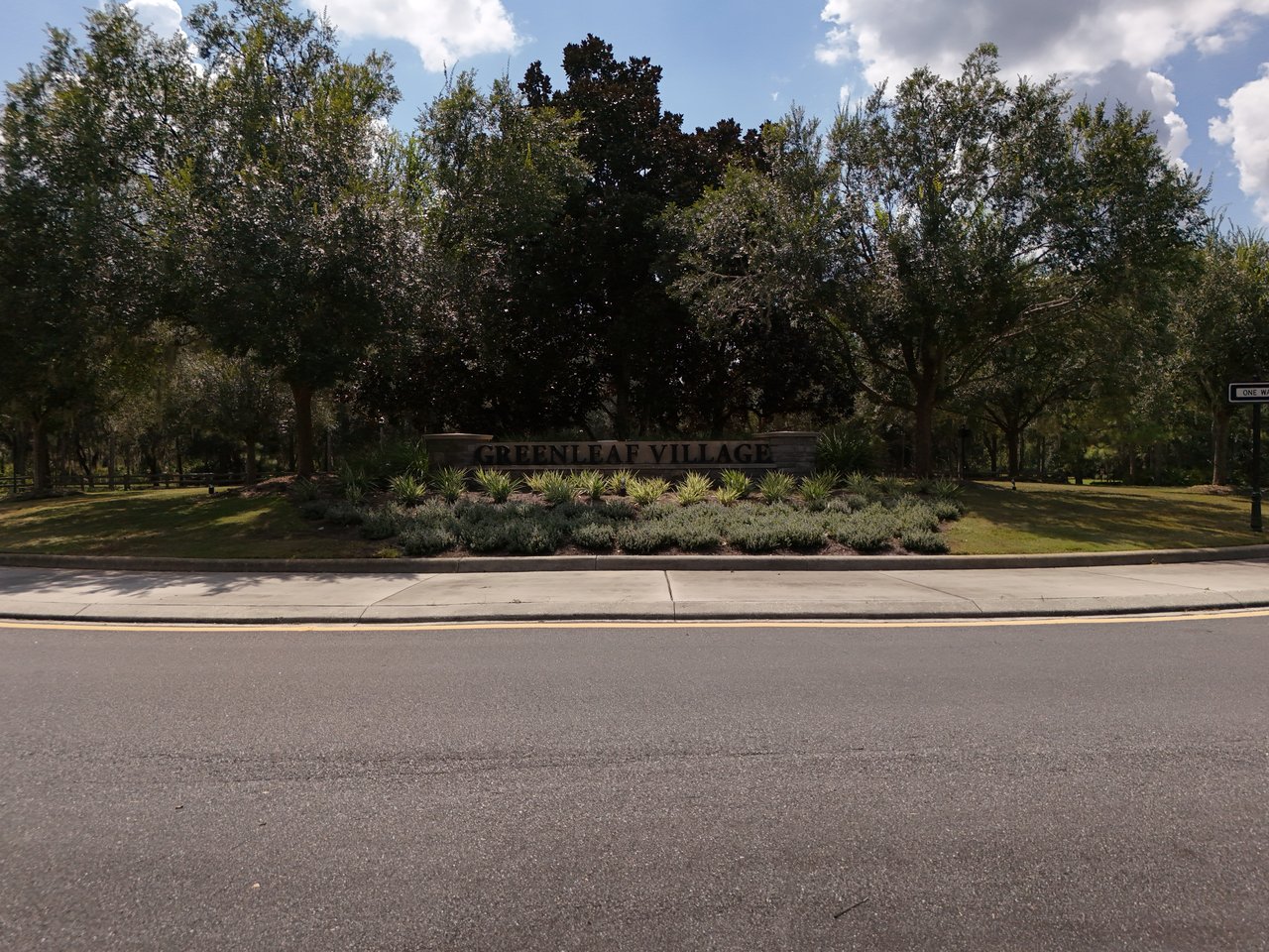 Greenleaf Village entrance sign amid landscaping and mature trees beside the road.