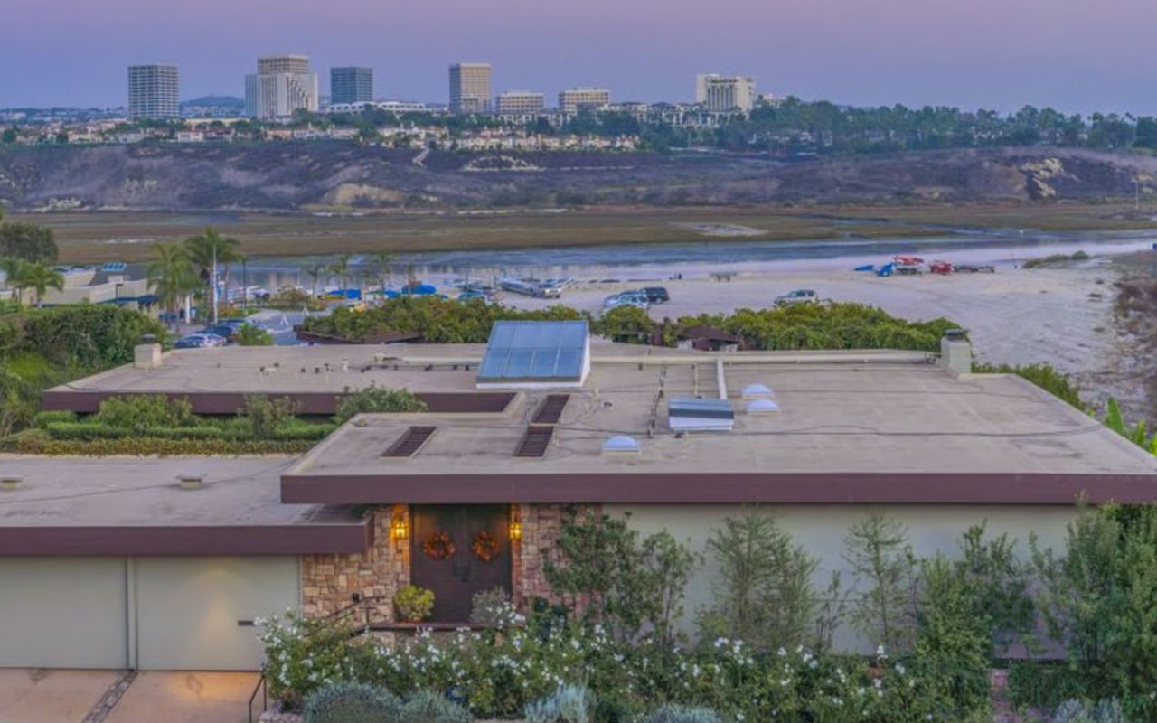 A two-story building with a flat roof, overlooking a sandy area and a city skyline in the distance.
