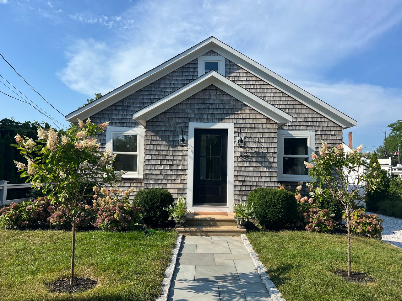 Gray cedar-shingle house with white trim, black front door, flowering shrubs, and a stone walkway.
