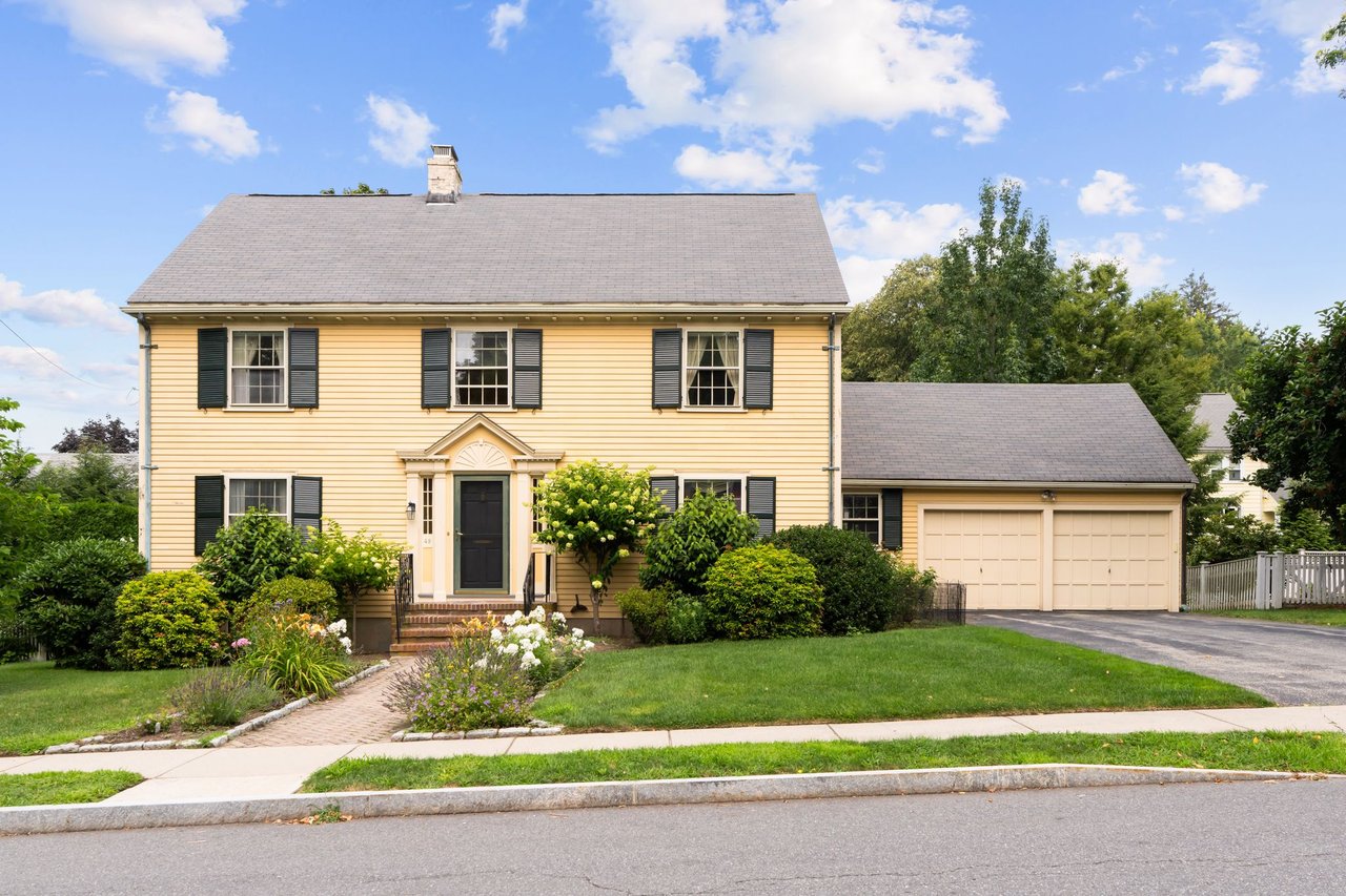 Yellow Colonial home with black shutters, an attached garage, and landscaped front yard.