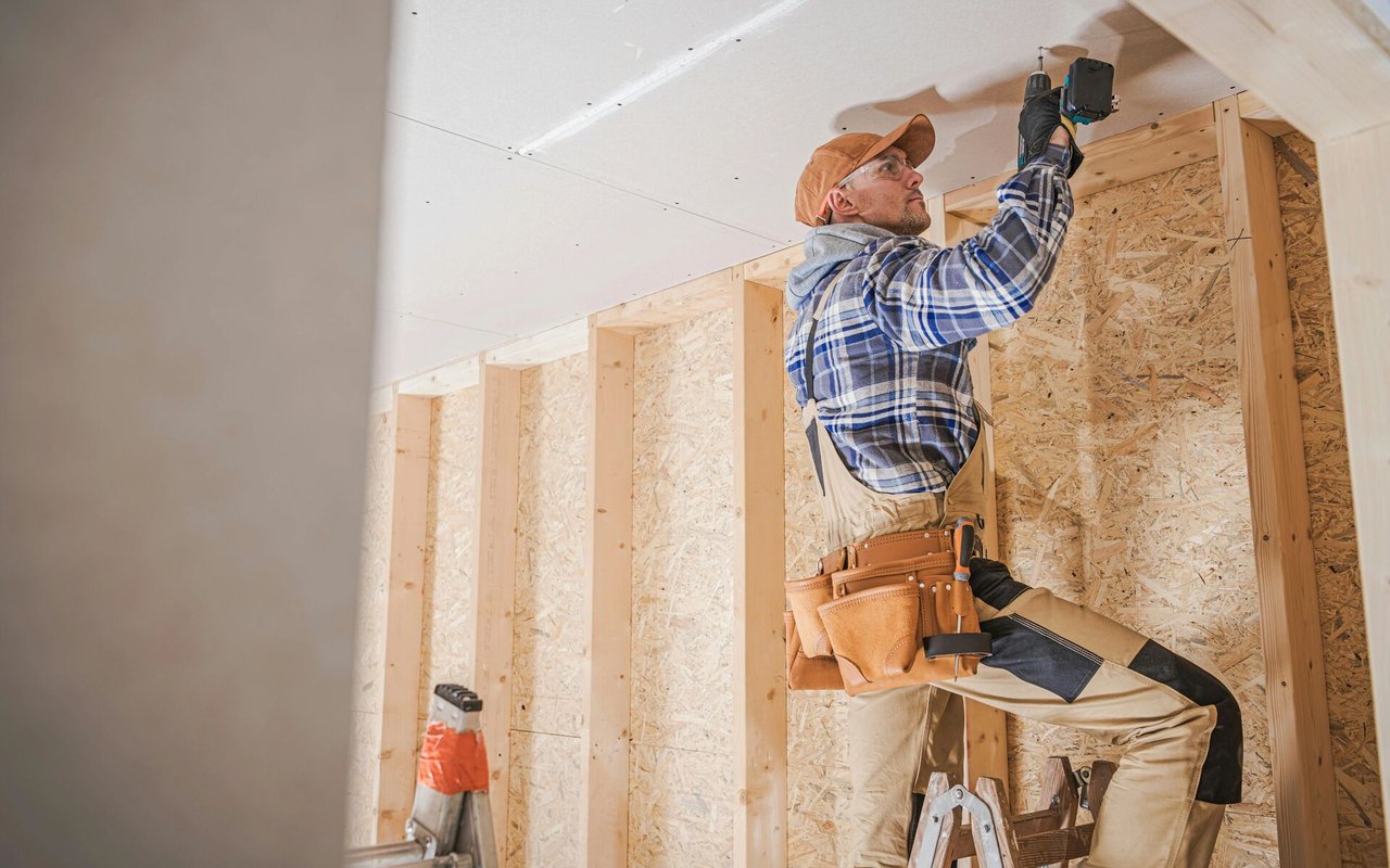 Worker fastening ceiling drywall in an unfinished room with exposed wood framing.