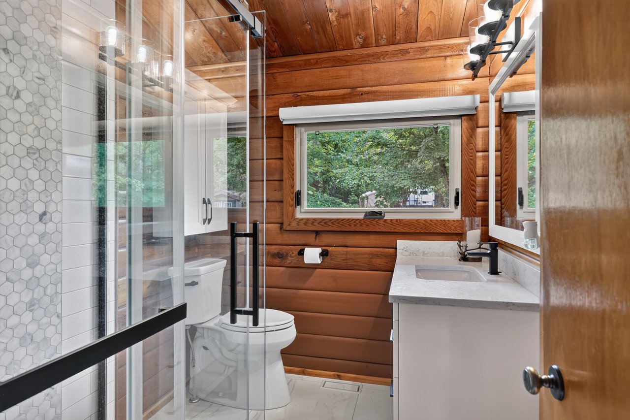 Bathroom with warm wood walls, a glass shower, and a white vanity beneath a large window.
