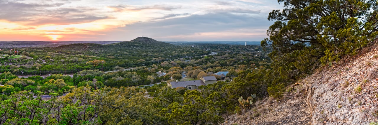 Wooded Hill Country hills and scattered homes beneath a colorful sunset.