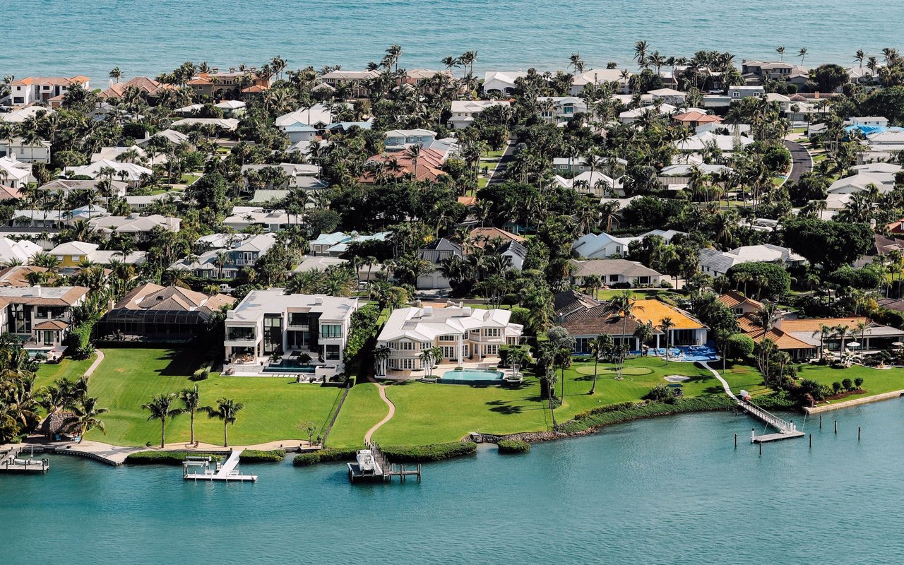 Aerial view of a waterfront neighborhood with palm-lined homes, lawns, private docks, and a pool.