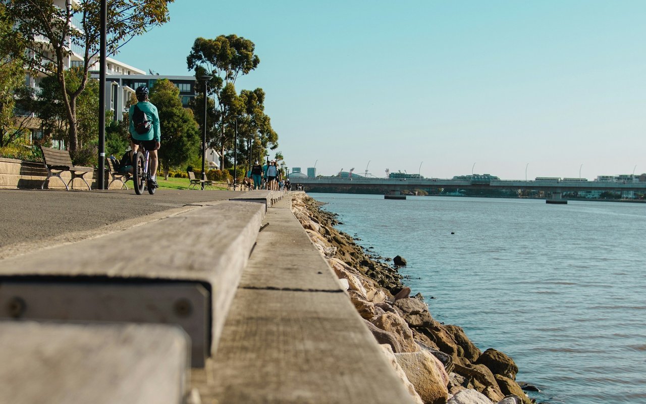 Waterfront promenade with a cyclist, benches, rocky shoreline, and a bridge in the distance.
