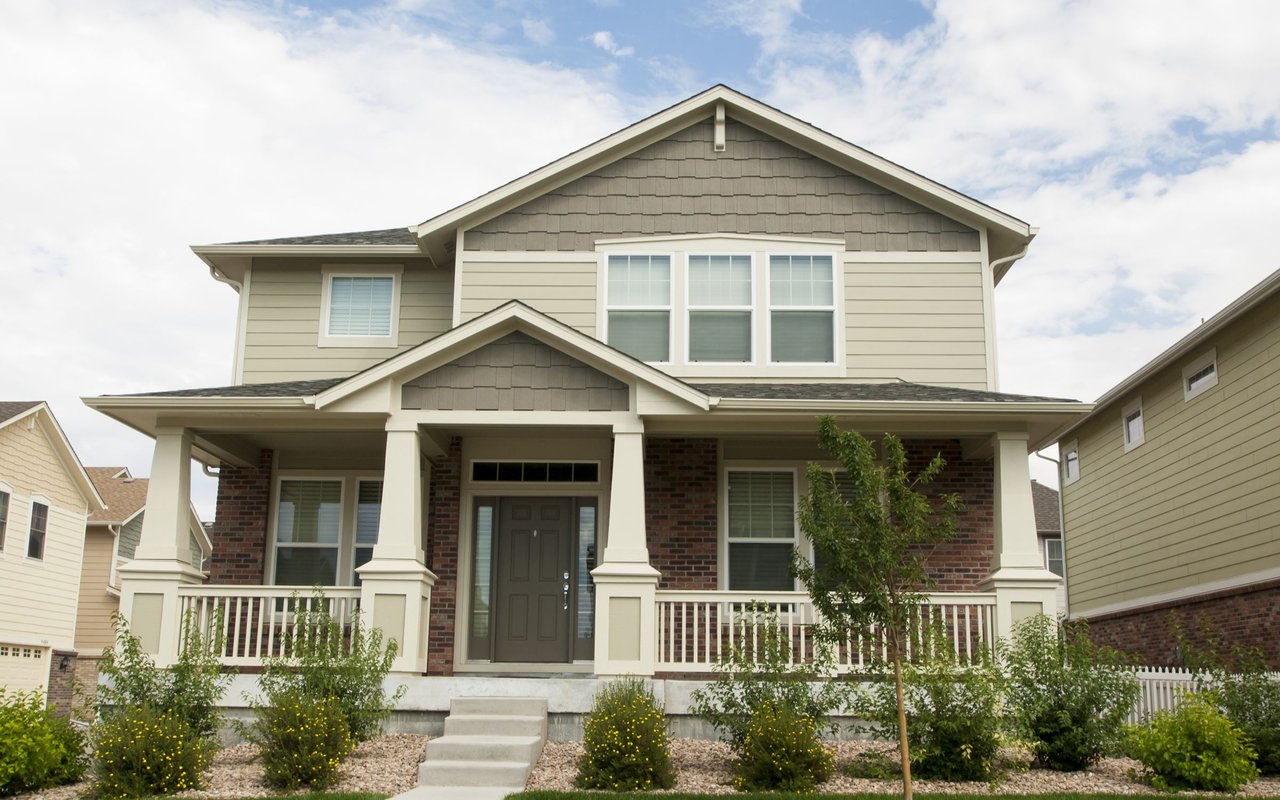 Two-story beige house with a brick facade, covered porch, and landscaped front yard.