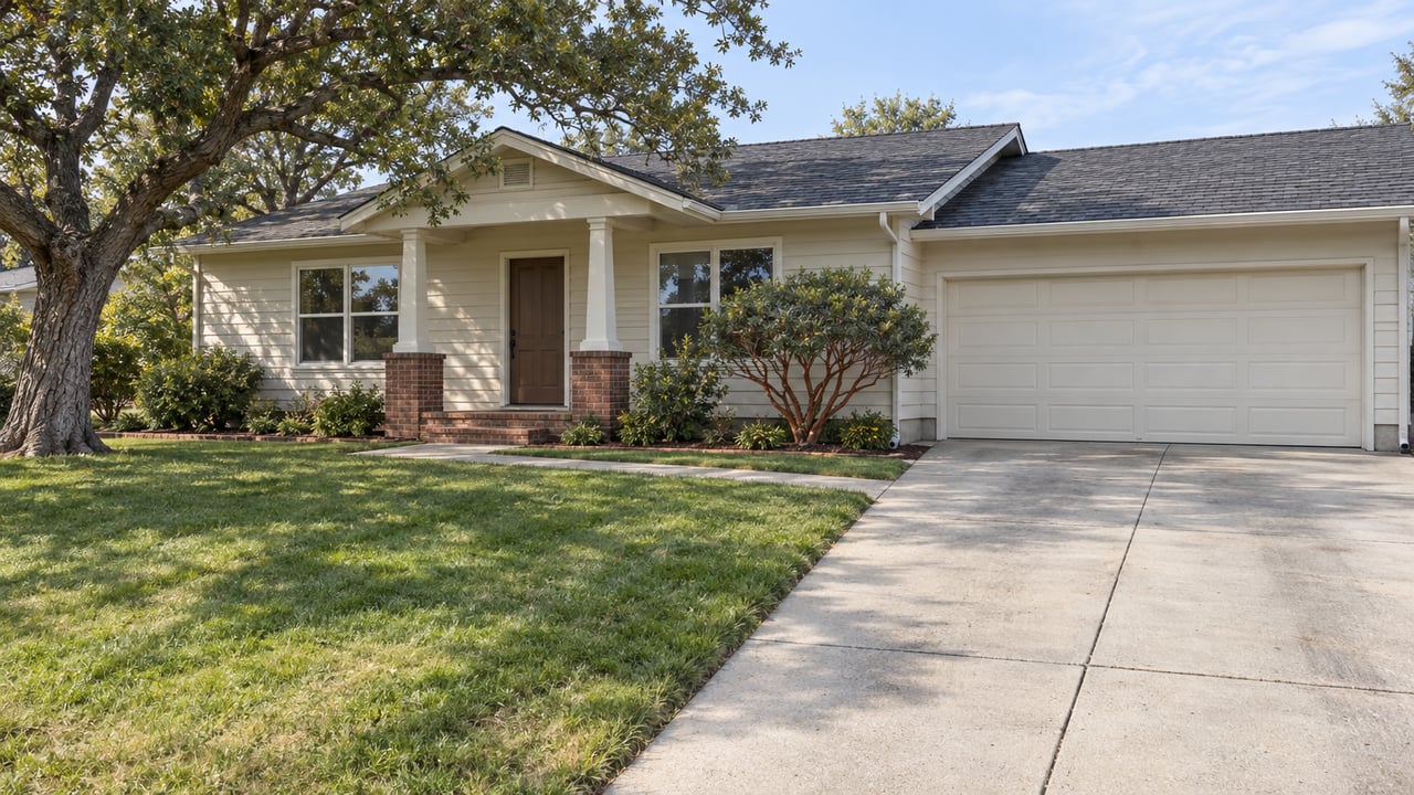 An ivory-sided single-story home with a brick porch, attached garage, deep front lawn and mature live oak in gentle late-morning light.