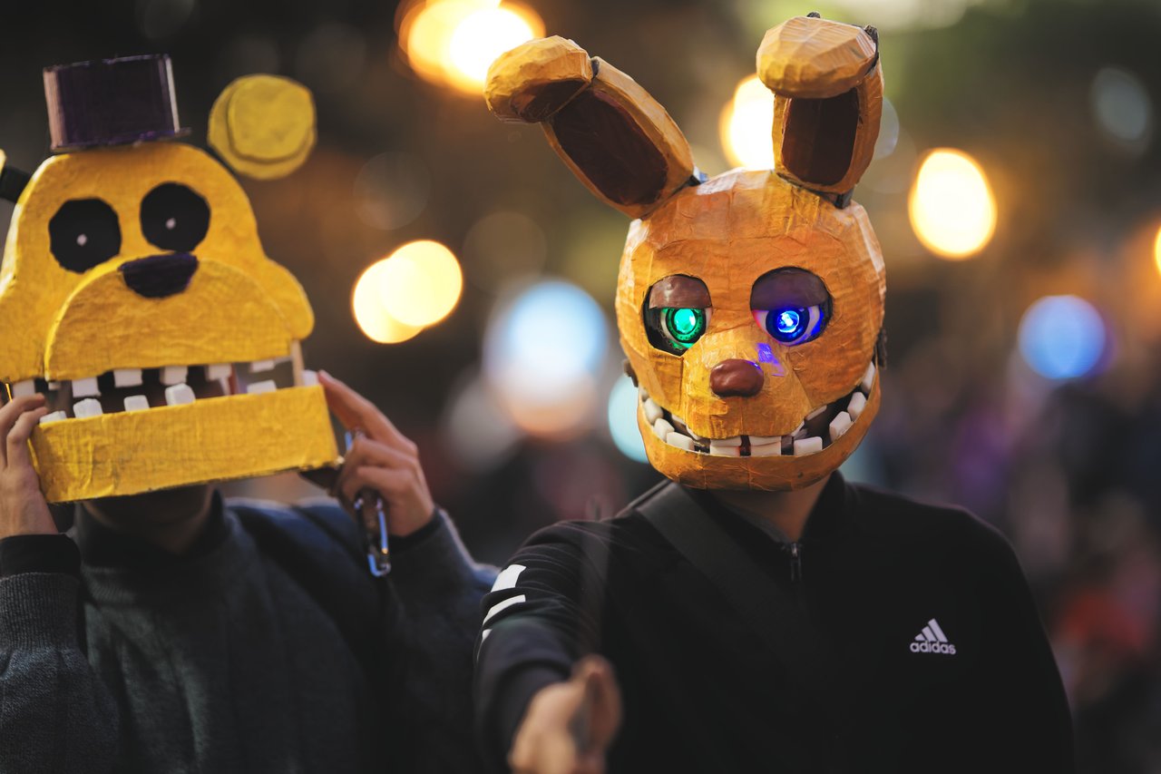 A vibrant nighttime photograph of the Greenwich Village Halloween Parade featuring elaborate costumes, illuminated giant puppets, crowds along Sixth Avenue, and historic Manhattan buildings decorated for Halloween.
