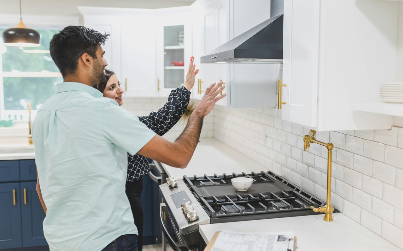 Modern kitchen with a couple examining the dark range hood and gas cooktop.