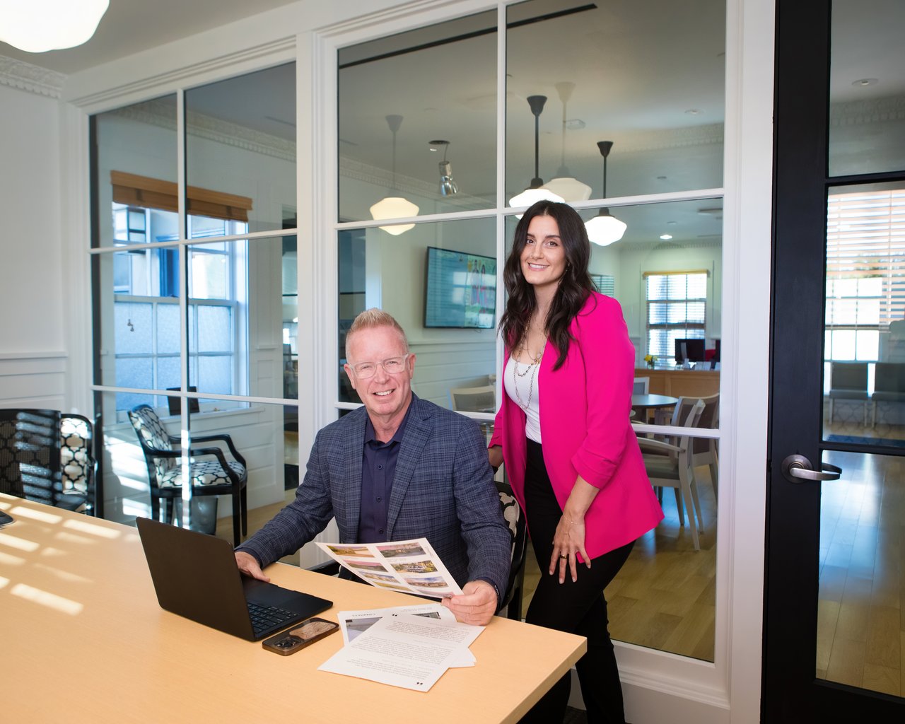 Two We Group members pose in their office, one seated with property brochures and a laptop.