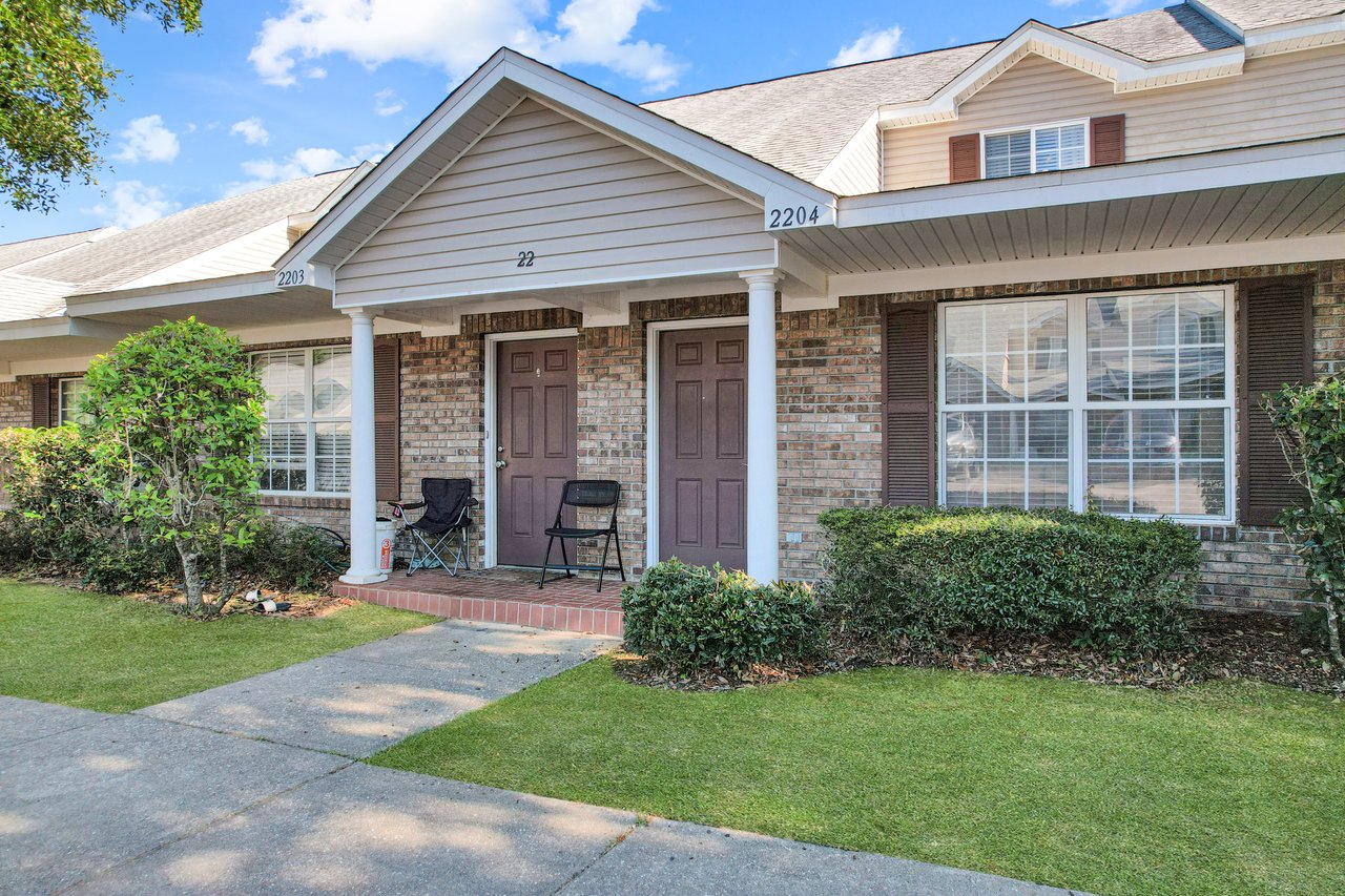 Suburban townhouse exterior with two brown doors and a brick facade. Two folding chairs and small table on porch, surrounded by shrubs and a manicured lawn.