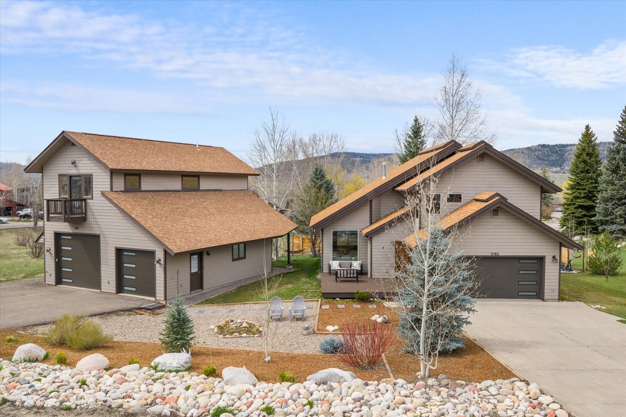 Two taupe mountain homes with multiple garages, landscaped rock gardens, and wooded hills behind them.