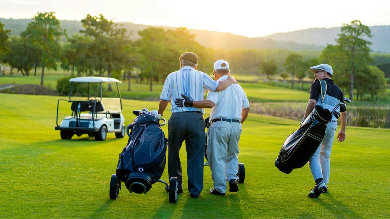 Three golfers walk across a green fairway at sunset with golf bags and a cart.