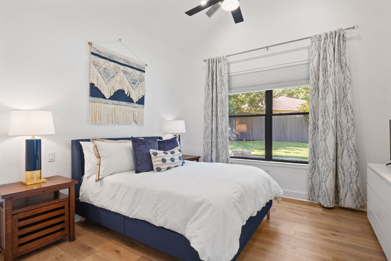 Furnished bedroom with a navy bed, patterned curtains, and a window overlooking a fenced yard.