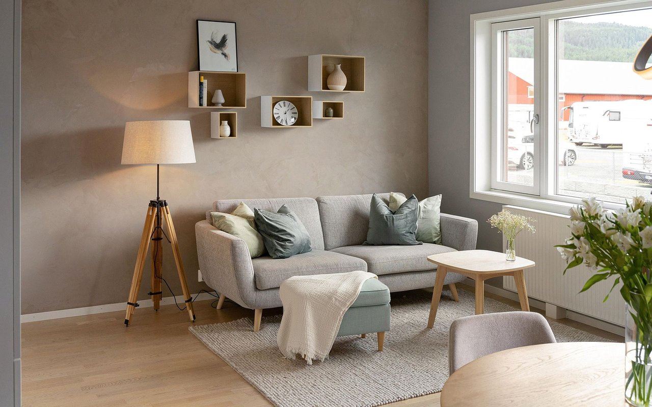 Staged living room with a gray sofa, tripod lamp, floating shelves, and a large daylight-filled window.