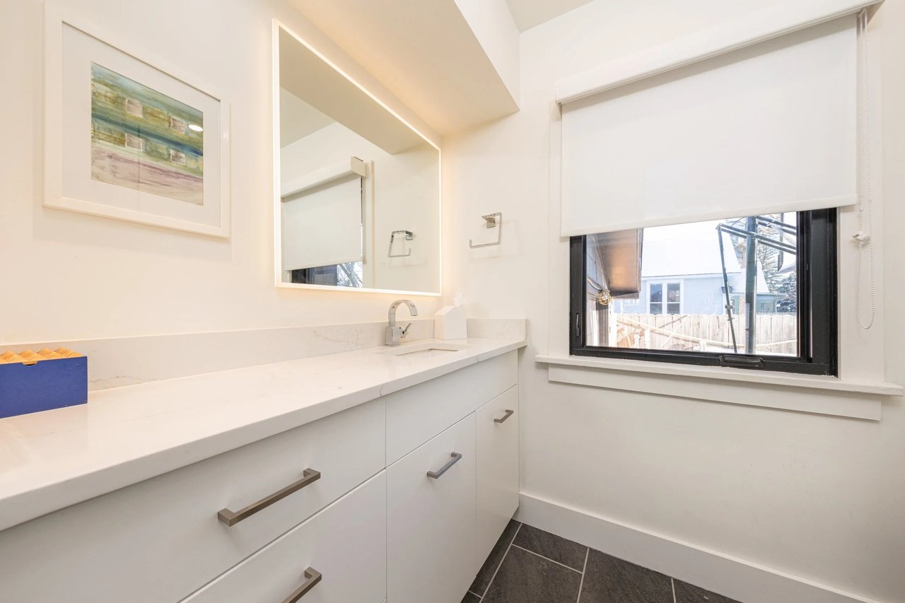 Renovated bathroom with a long white vanity, illuminated mirror, and black-framed window.