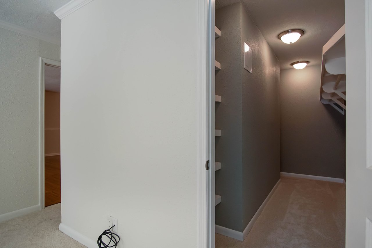 Dimly lit hallway with beige walls and carpet, featuring two ceiling lights. A partially visible room on the left and coiled cables on the floor.