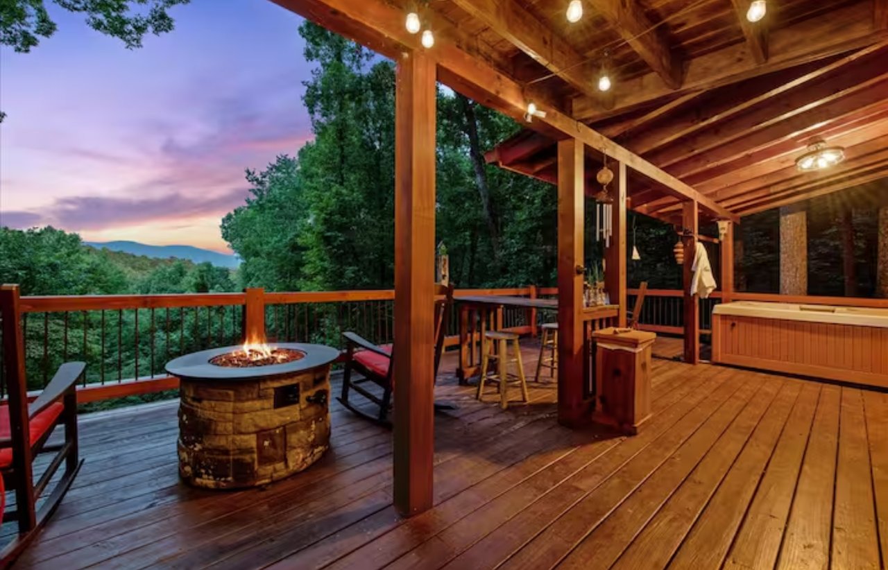 Covered wooden deck with a stone fire pit, hot tub, and wooded mountain views at dusk