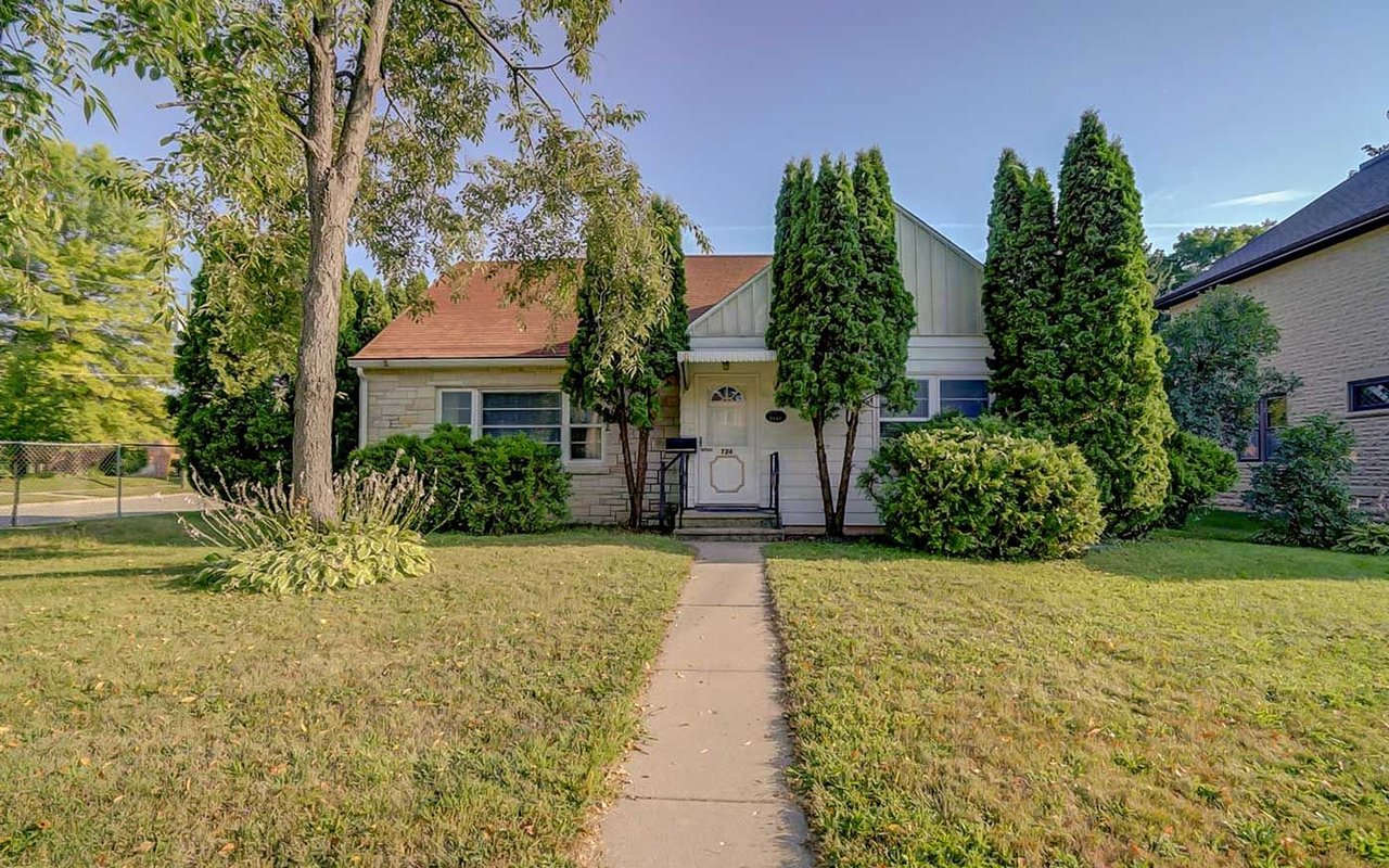 Single-story stone house with a front walkway, red-brown roof, and tall evergreen shrubs.