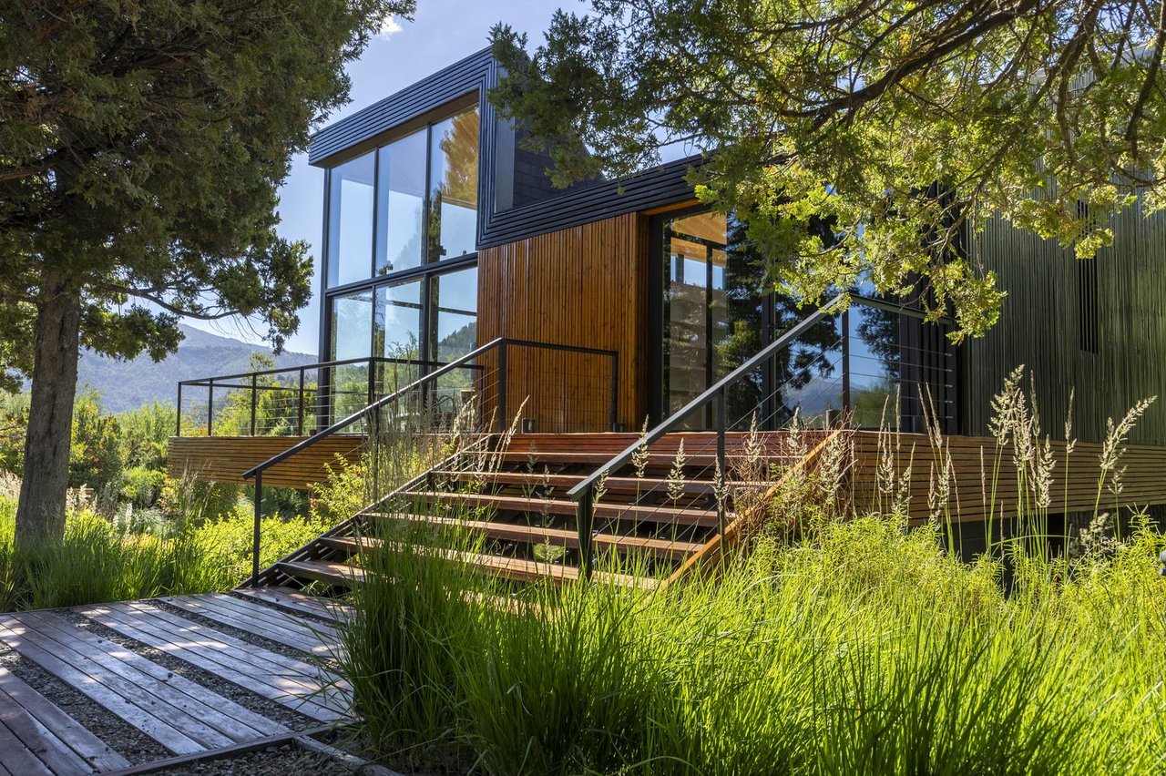 Modern glass-and-wood house with a broad timber deck, surrounded by trees and tall grasses.