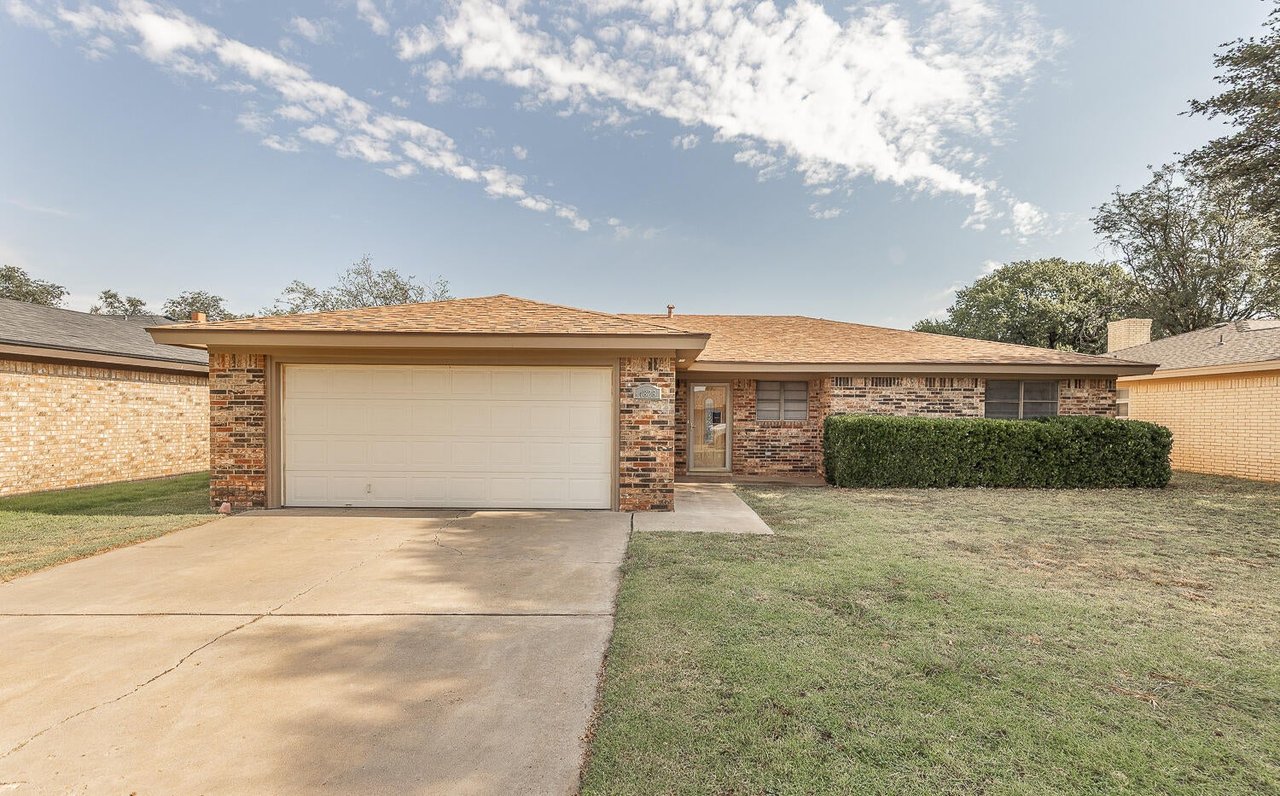 Single-story brick home with a wide garage, concrete driveway, and trimmed front hedge.