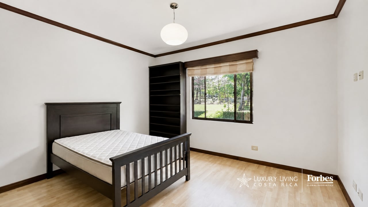 Bedroom with a dark wood bed, tall bookshelf, and window overlooking greenery.