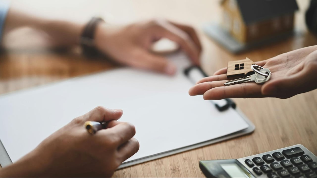 Hands exchange a house key above mortgage paperwork and a calculator.