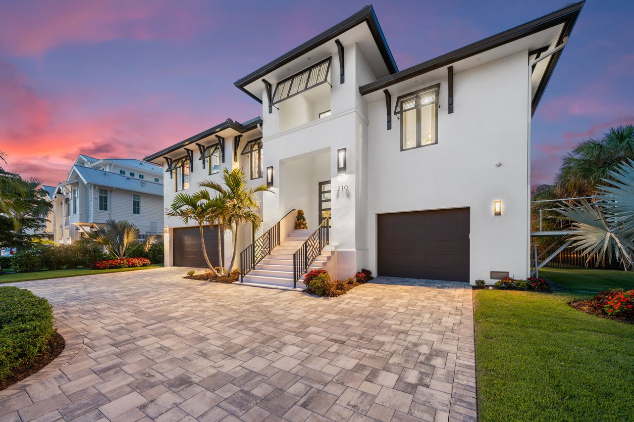 Modern white two-story home with angular black rooflines and a broad paver driveway at dusk.