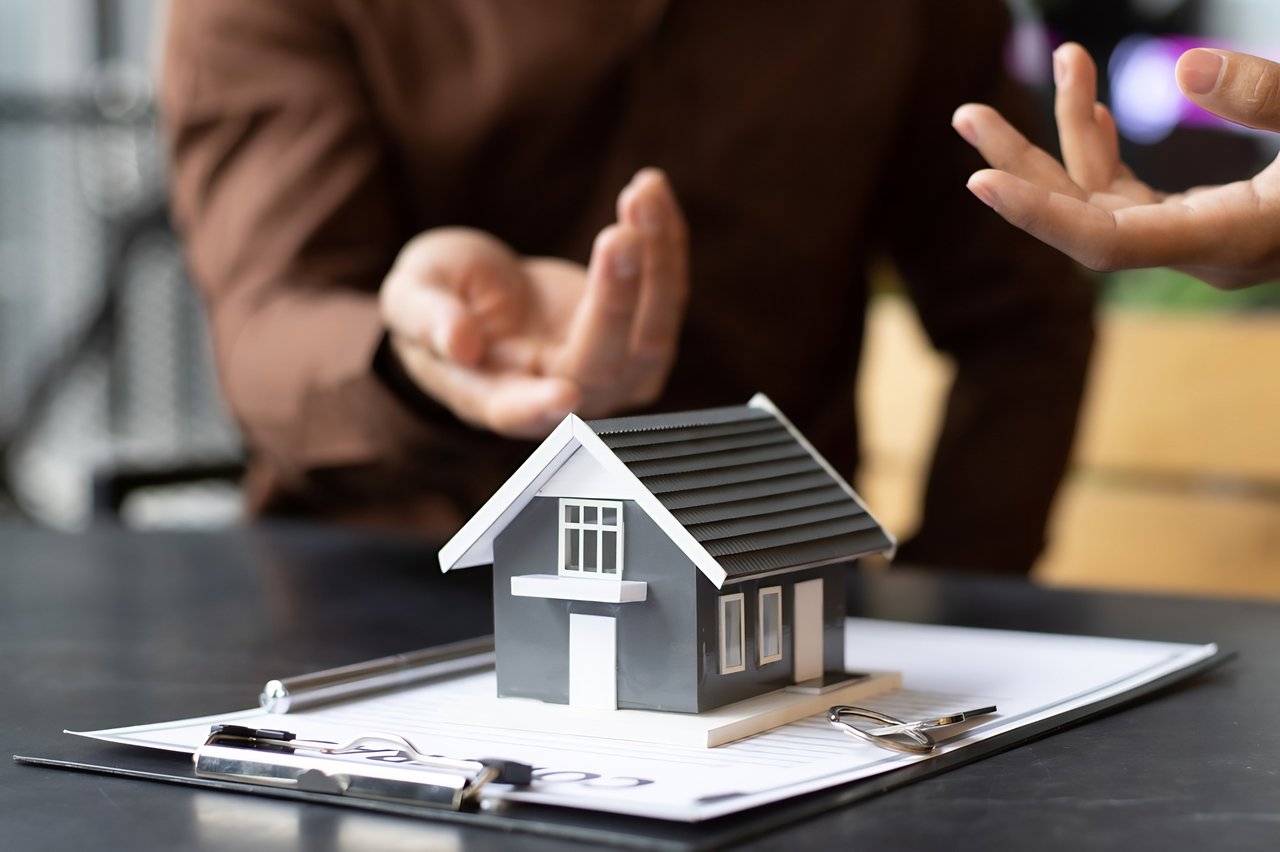 Miniature gray house on paperwork as people gesture during a real estate discussion.