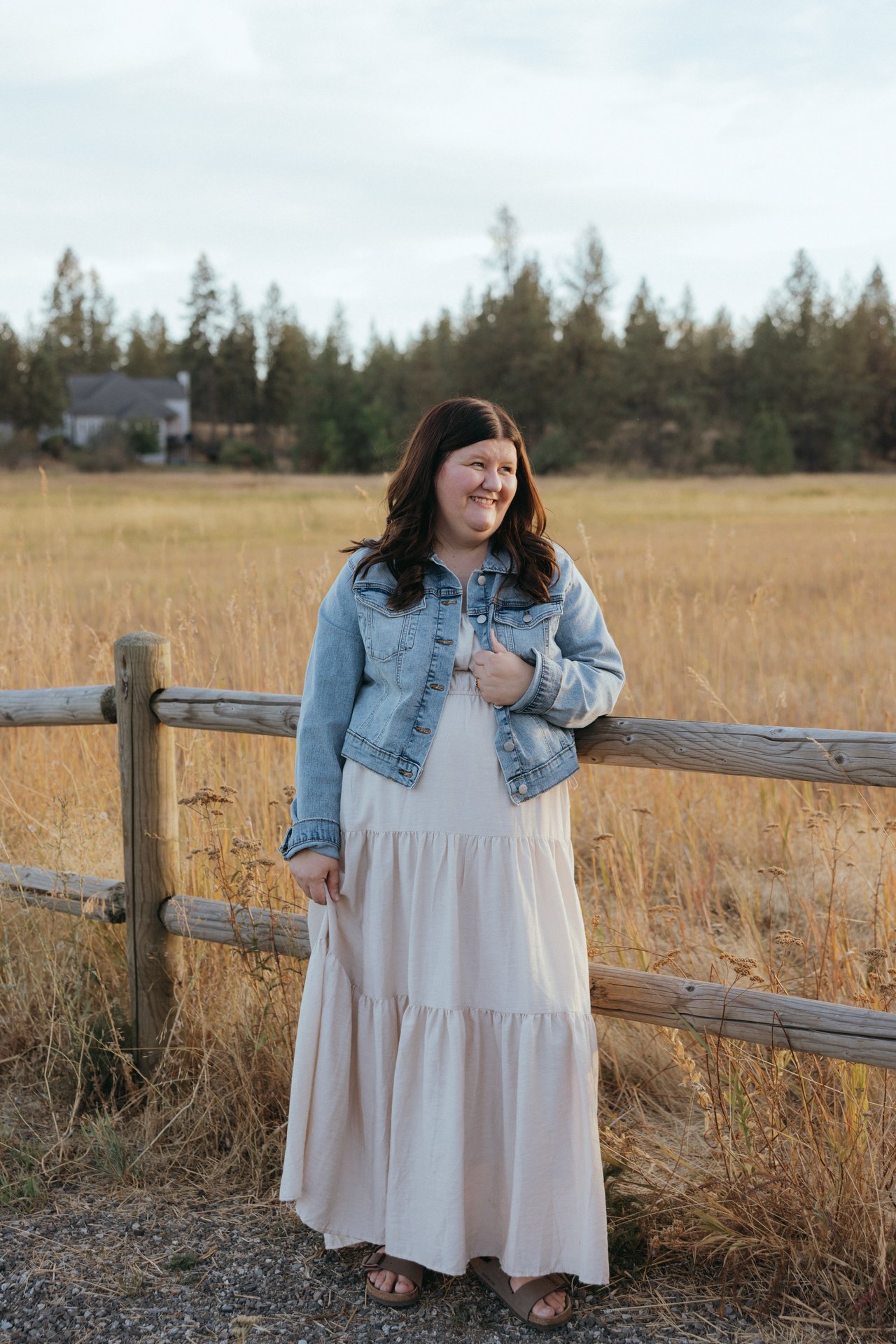 Smiling woman in a denim jacket and pale dress standing by a wooden fence in a field.